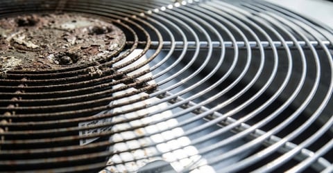 Close-up of an air conditioning units fan grille showing curved metal fins with dirt and dust accumulation on one side