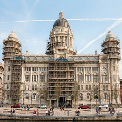 Historic stone government building with large dome and multiple towers under renovation, waterfront location with pedestrians in foreground