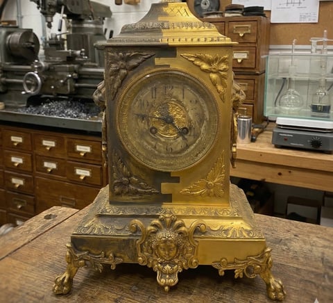 Ornate brass mantel clock with decorative eagle relief and Roman numerals, displayed on wooden workbench in workshop setting
