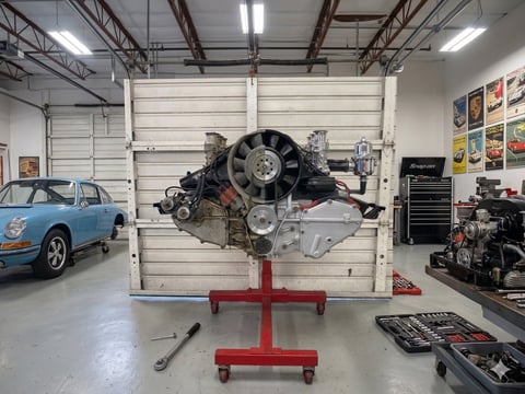 Engine mounted on red stand in automotive workshop with blue car and tools visible