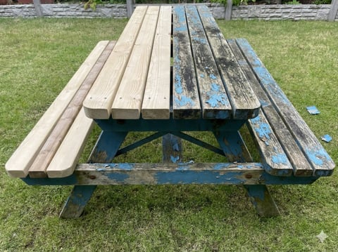 Weathered wooden picnic table with blue painted base on grass, showing worn bench seating and natural wood aging