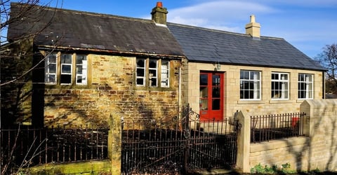 Stone cottage with cream and yellow rendered walls, black roof, red door, white-framed windows, and black iron fence in sunny countryside setting