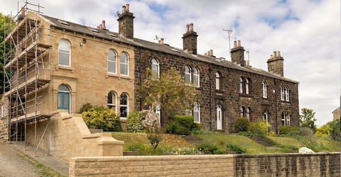Historic stone building with ivy, chimneys, and scaffolding on left side, situated on a landscaped hillside