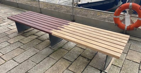 Wooden slatted bench on a paved dock with metal legs, life ring and railing visible in background